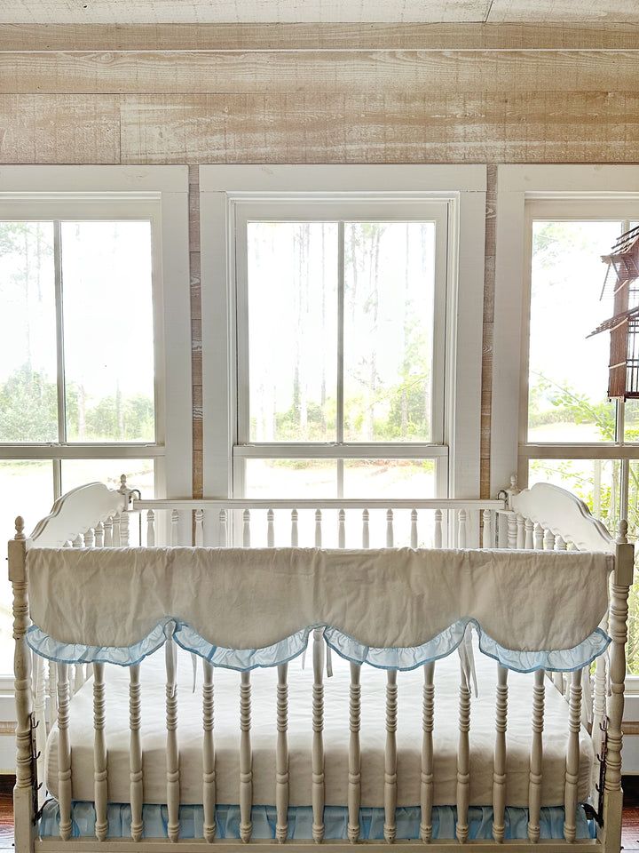 Scalloped rail cover on a white crib framed by tall windows and pine woods beyond