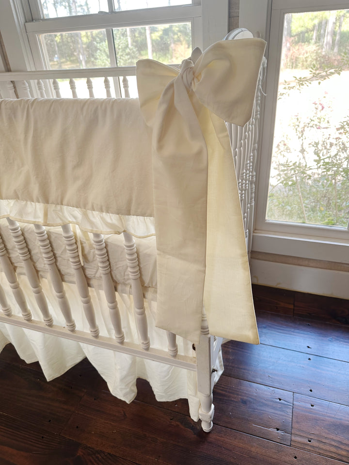 Ivory bow with long sash tails tied at the crib post next to a sunny window, rail cover ruffle and ties visible
