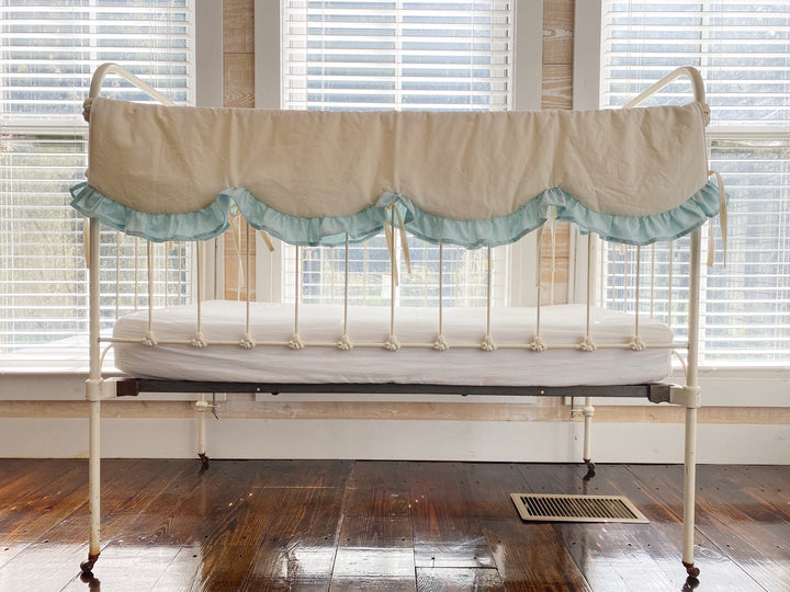 Ivory scalloped crib rail cover with a soft mist blue ruffle on an antique white iron crib