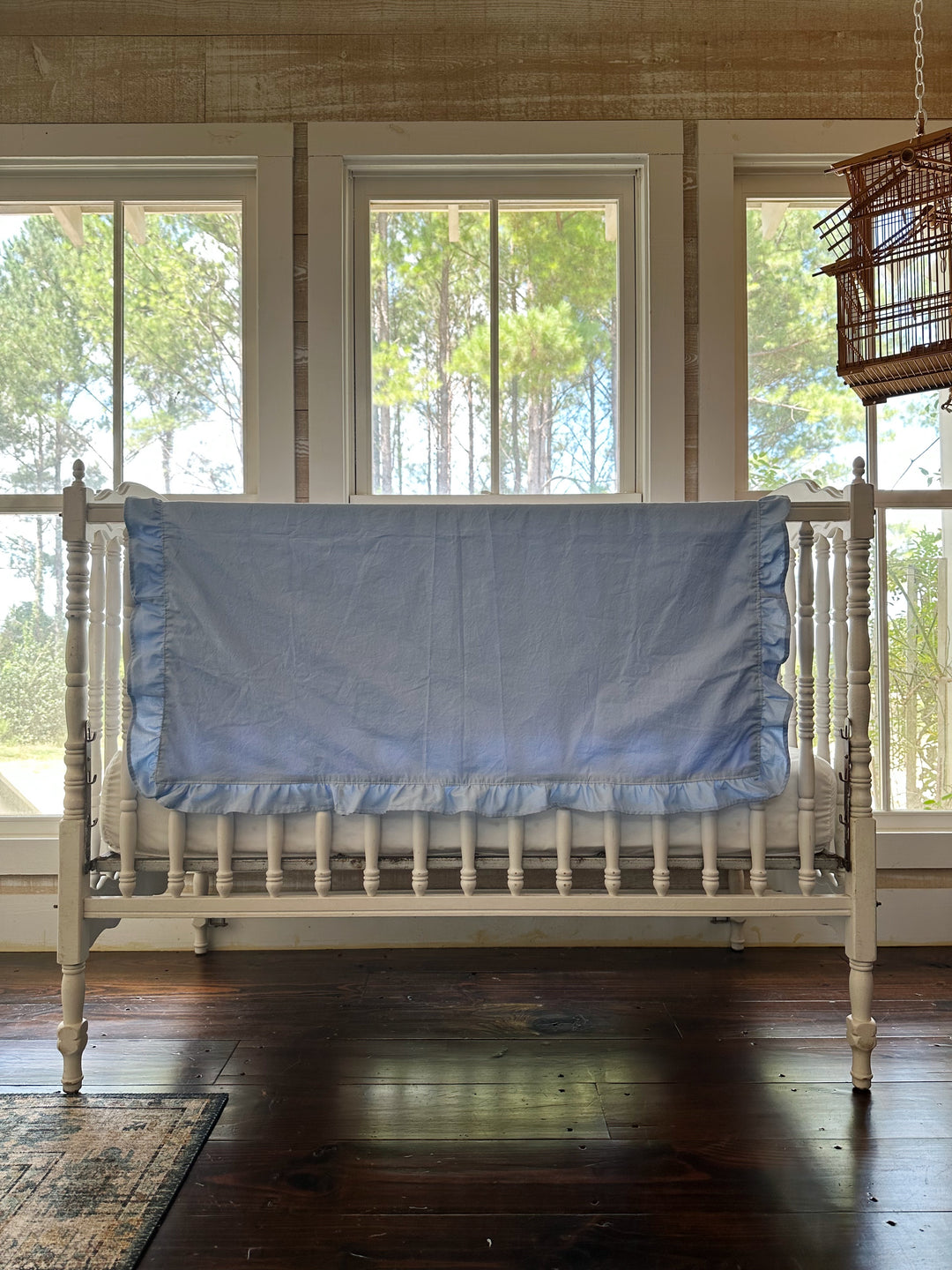 Wide nursery view of a baby blue ruffled crib blanket draped across a white crib rail above a hardwood floor - High Cotton Textile