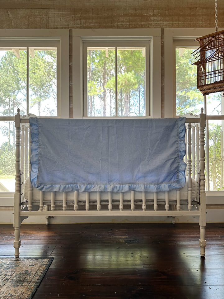 Wide nursery view of a baby blue ruffled crib blanket draped across a white crib rail above a hardwood floor - High Cotton Textile
