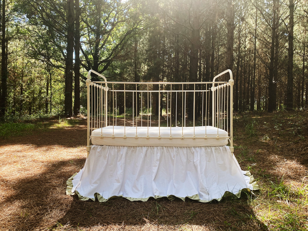 White ruffled crib skirt with sage green ruffle trim on iron crib, end view showing 22 inch drop in forest setting