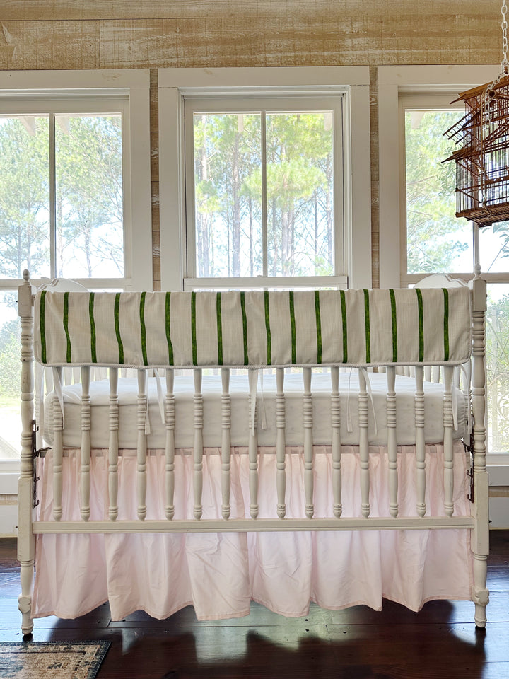 Green and white striped tailored crib rail cover above a gathered baby pink farmhouse crib skirt on a white spindle crib