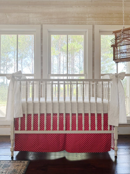 Cherry red and white polka dot tailored crib skirt with two large white bows tied to the front posts of a white spindle crib