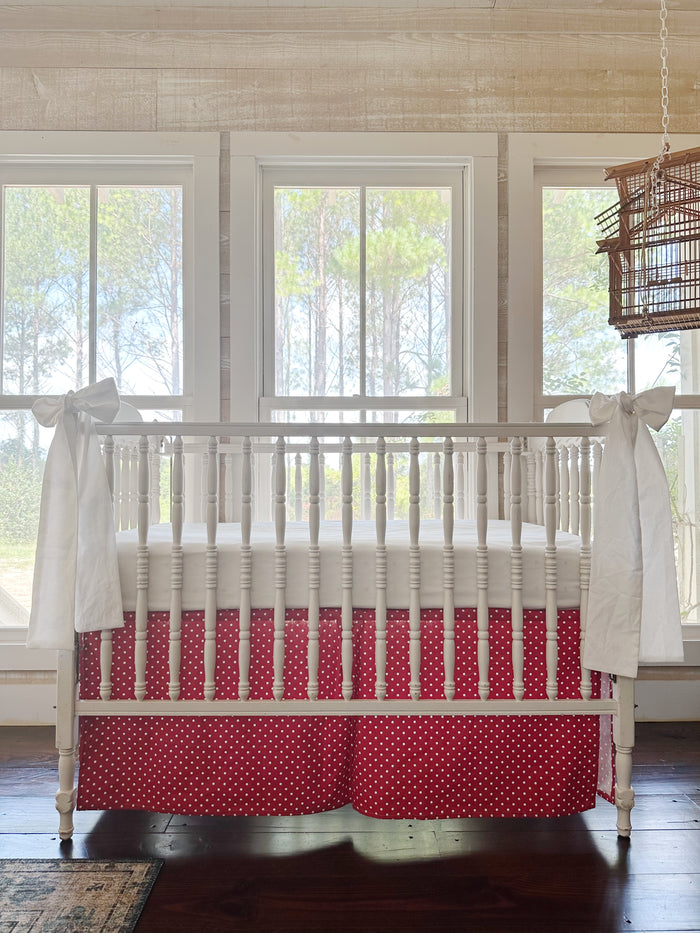 Cherry red and white polka dot tailored crib skirt with two large white bows tied to the front posts of a white spindle crib