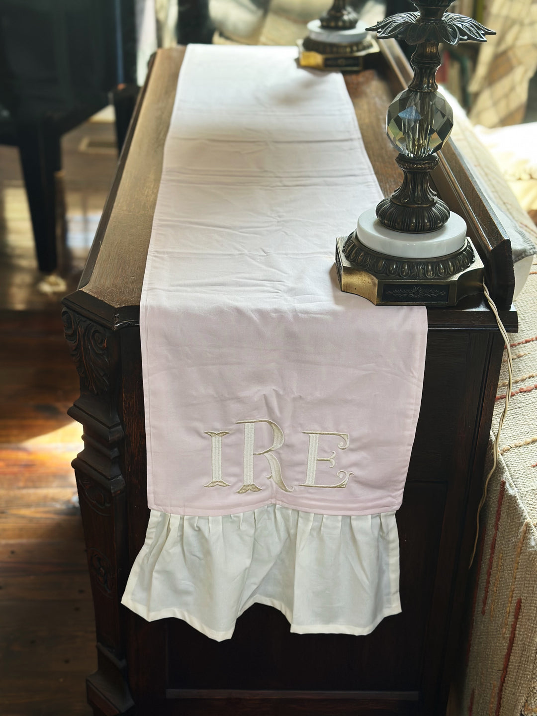 Baby pink dresser runner with an ivory ruffle on a carved antique dresser, the monogrammed end hanging over the side beside a brass and crystal lamp