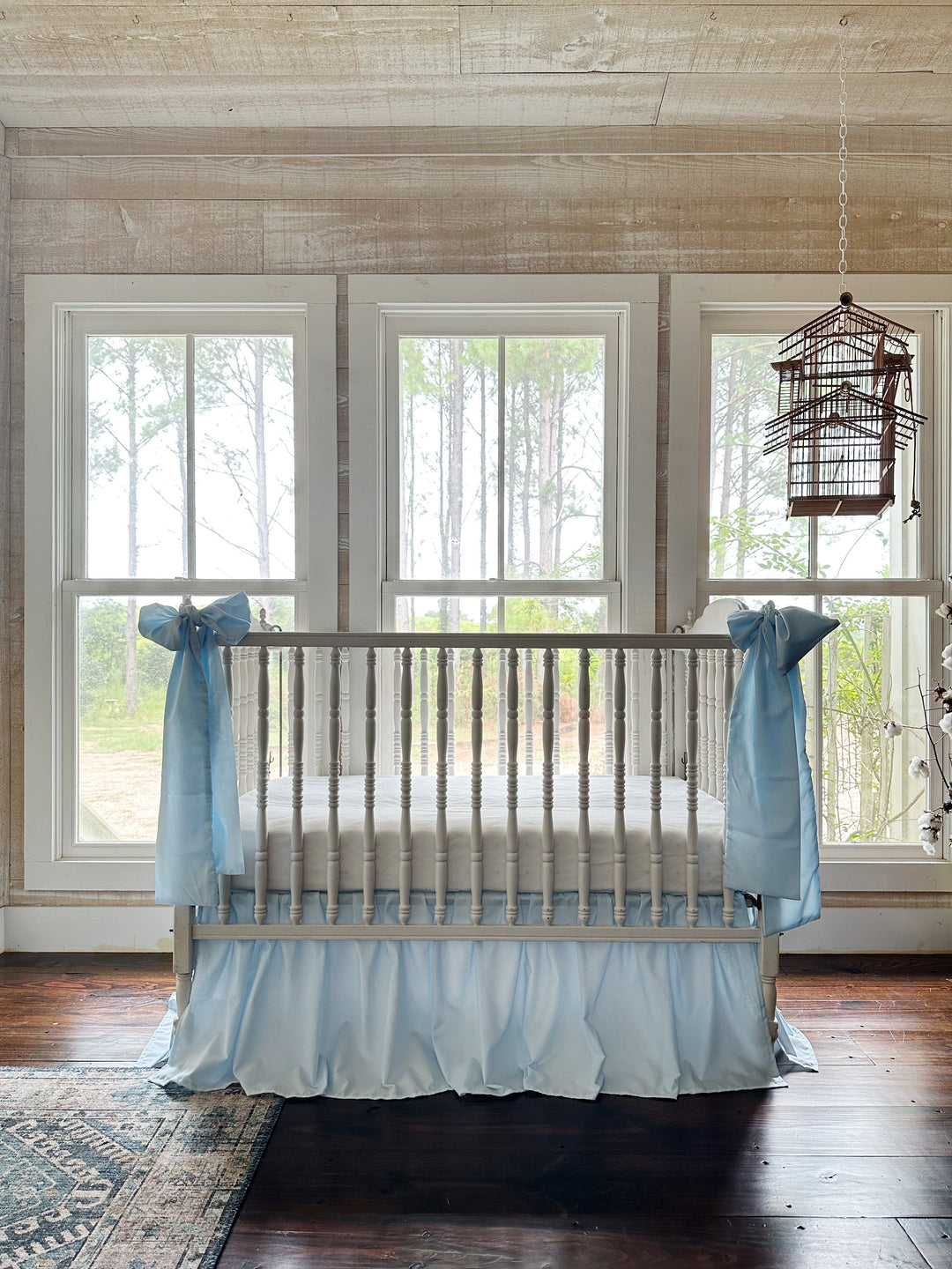 Heirloom blue crib skirt and bows in a nursery beneath three windows framed by pines