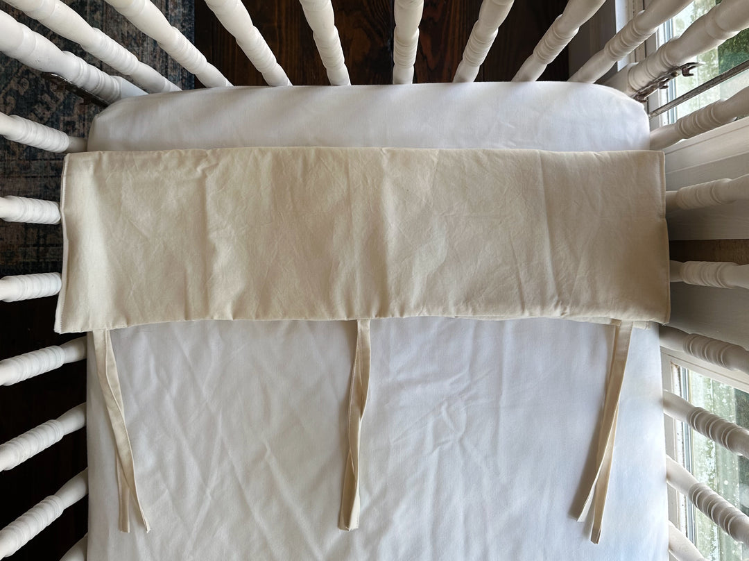 Folded Natural crib rail cover on a crib mattress showing three evenly spaced cotton ties