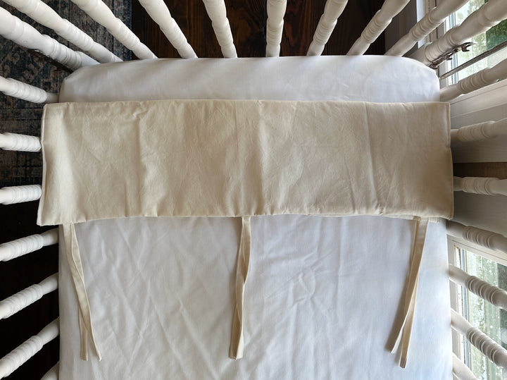 Folded Natural crib rail cover on a crib mattress showing three evenly spaced cotton ties