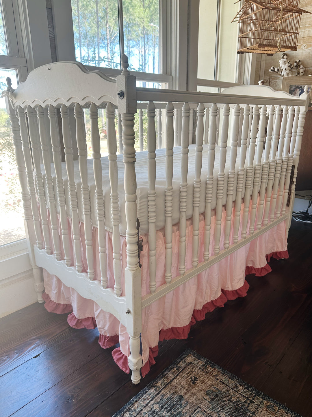 Three-quarter view of the pink ruffled crib skirt wrapping the foot end of a white spindle crib beside the windows
