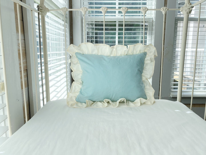 Ivory ruffled crib pillow in mist blue shown inside a white iron crib