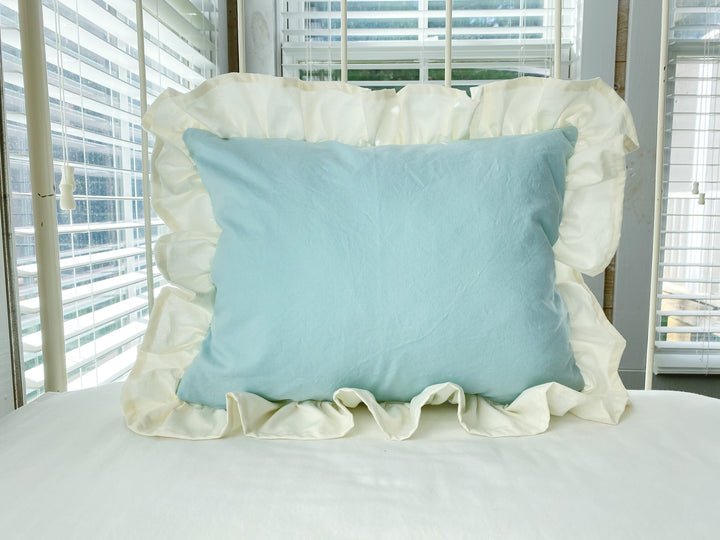 Mist blue crib pillow with a gathered ivory ruffle border resting on a white crib mattress