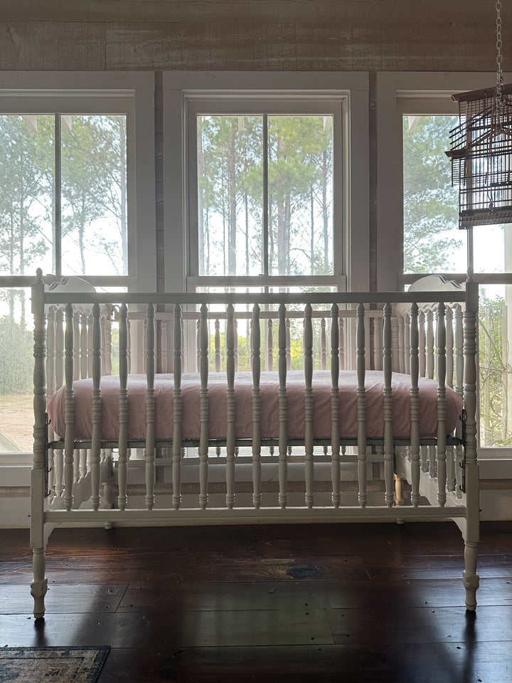 Front view of a white spindle crib with the Sisters Pink fitted crib sheet, pine trees through the nursery windows