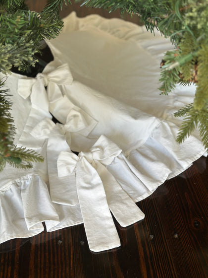 White ruffled Christmas tree skirt under a green tree, three white cotton bows tied down the front opening