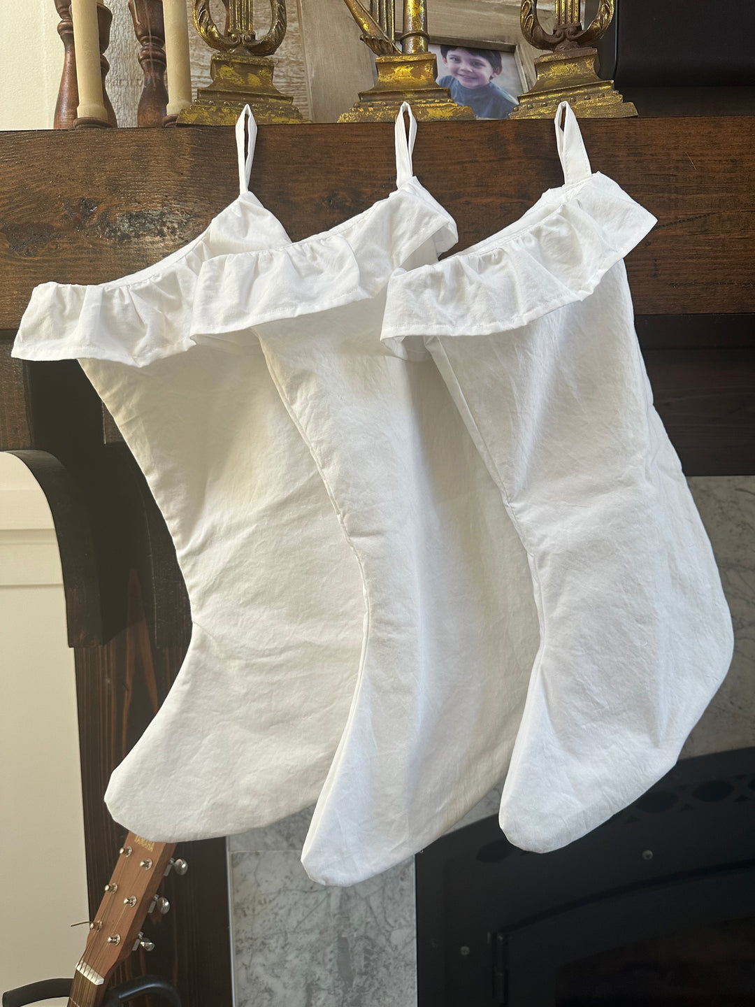 Three white farmhouse ruffled Christmas stockings hanging from a dark wood mantel below brass candlesticks