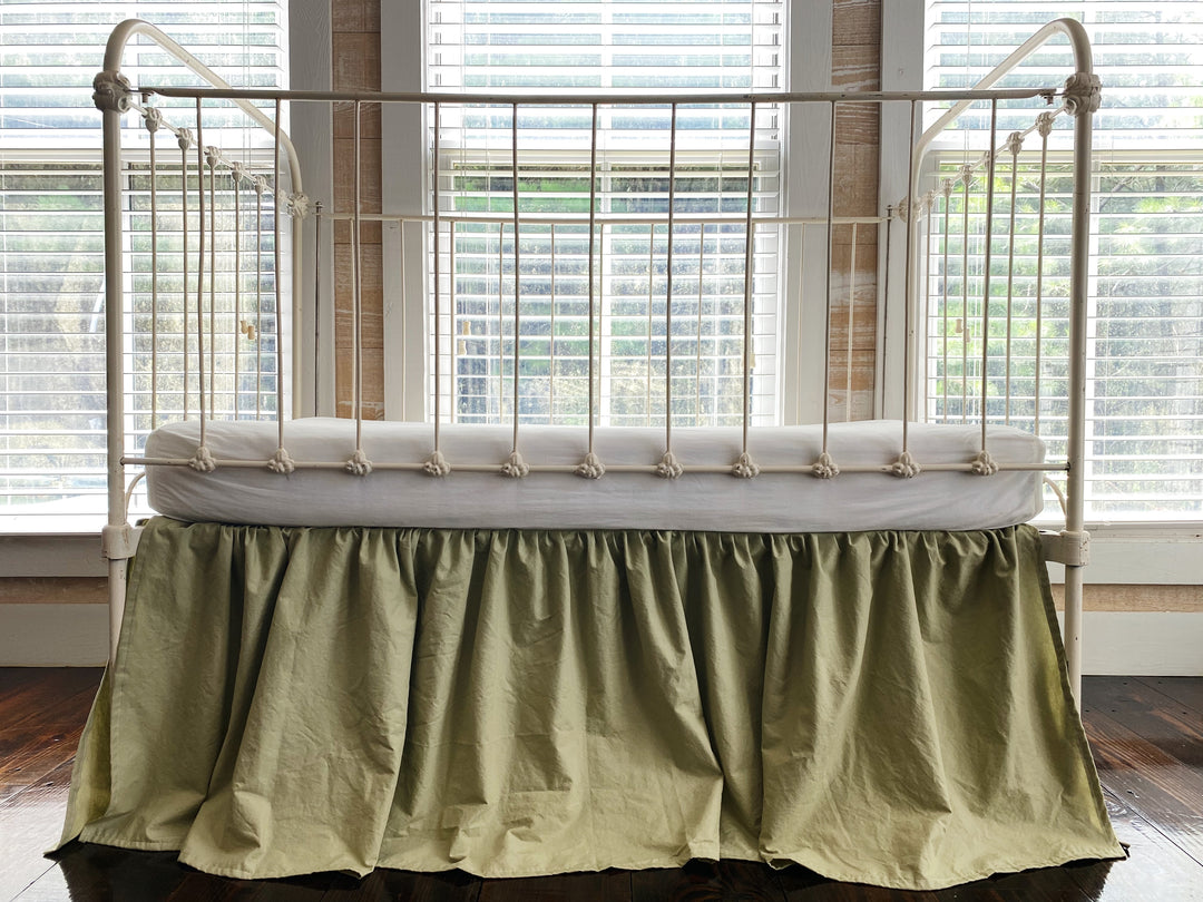 Floor-level close-up of sage green washed cotton crib skirt hem and split corner detail on hardwood floor