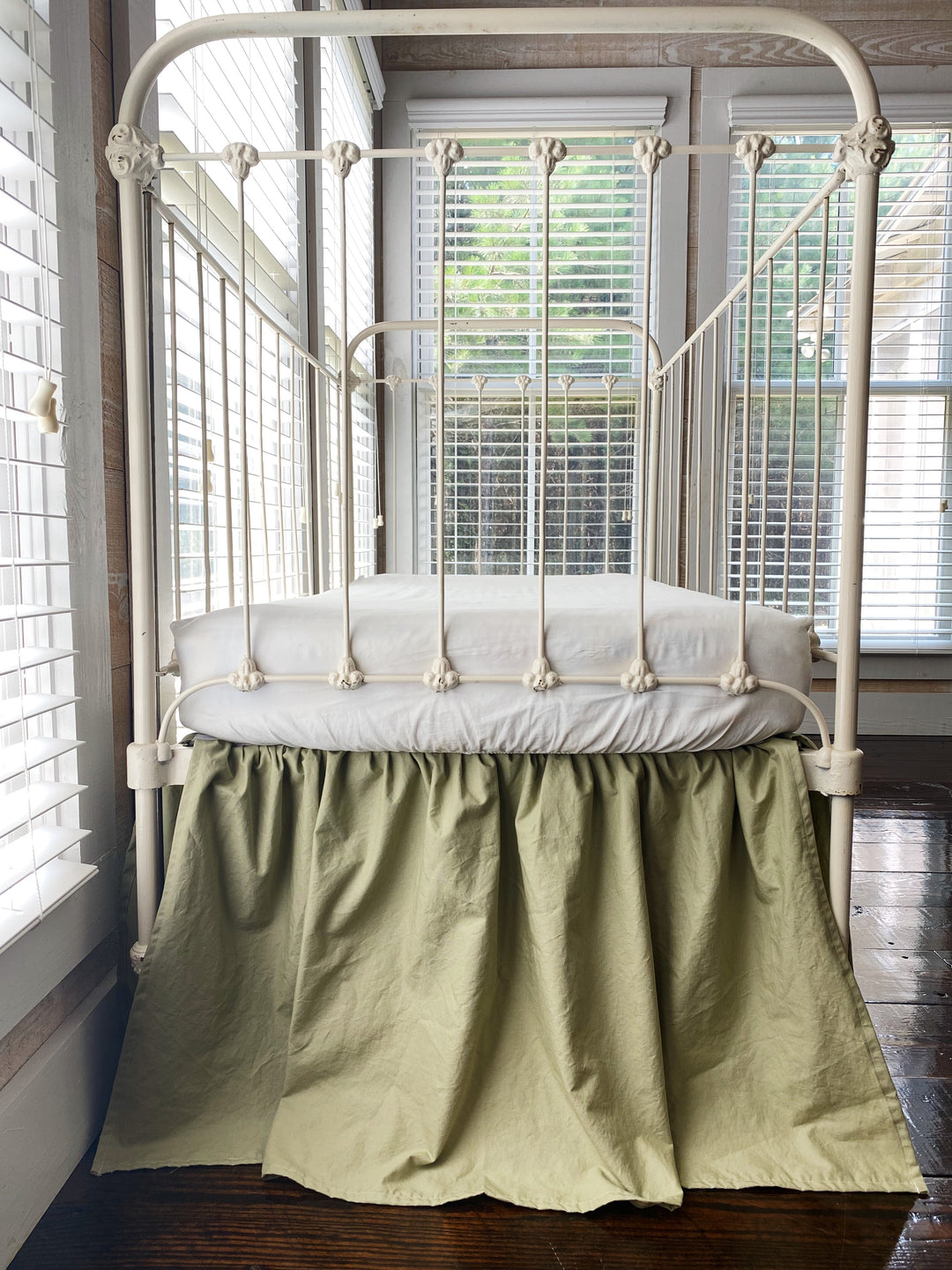 Sage green ruffled crib skirt on white iron crib, angled end view showing gathered silhouette and 20 inch length