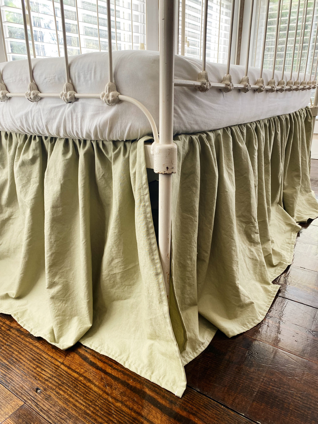 Sage green farmhouse crib skirt on iron crib, three-quarter angle view showing all-sided coverage and split corners