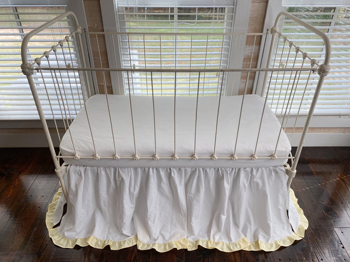 Overhead view of white gathered crib skirt with yellow ruffle trim and white fitted sheet on iron crib