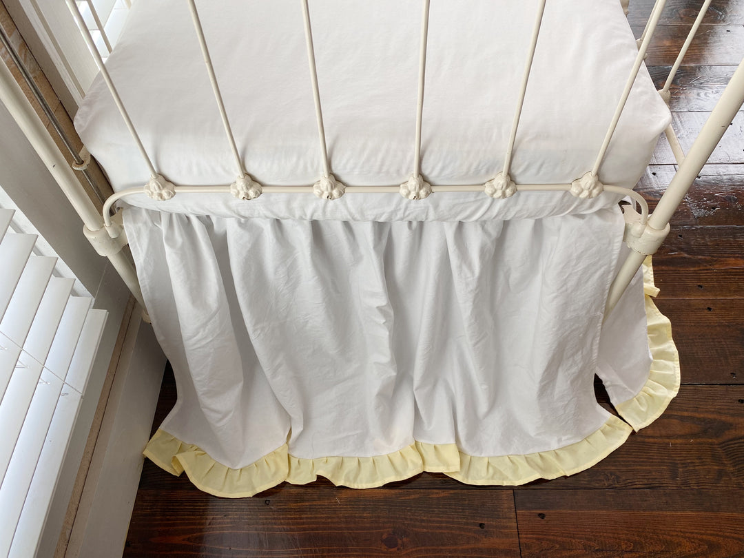 Top-down end view of white gathered crib skirt with baby yellow ruffle hem on iron crib, split corner visible