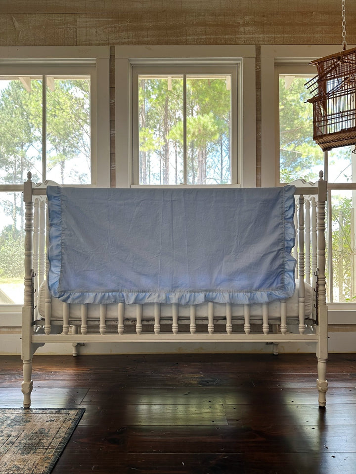 Wide nursery view of a baby blue ruffled crib blanket draped across a white crib rail above a hardwood floor - High Cotton Textile