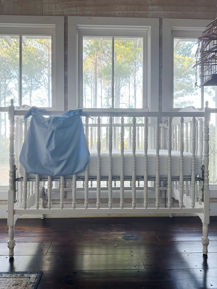 Baby blue crib caddy hanging on the outside of a white spindle crib in front of tall windows and pine trees