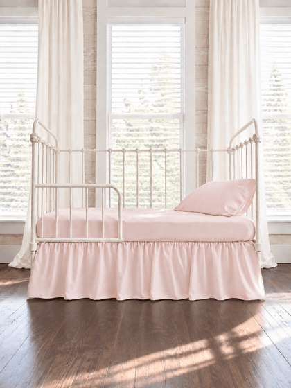 Baby pink farmhouse toddler bed skirt gathered on all four sides of a white iron crib converted to a toddler bed, with matching baby pink sheet and pillowcase - High Cotton Textile