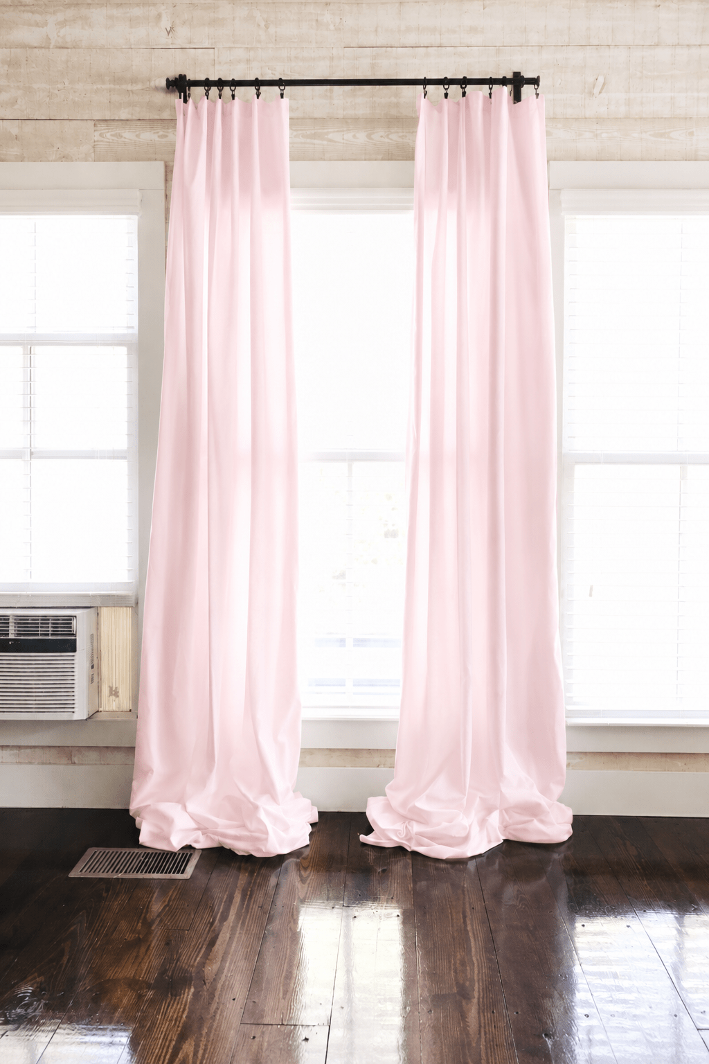Baby pink farmhouse nursery curtains hung as two panels on a black rod, puddling on dark wood floors between white-trimmed windows - High Cotton Textile