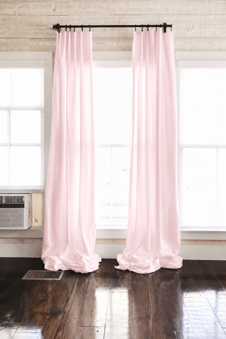 Baby pink farmhouse nursery curtains hung as two panels on a black rod, puddling on dark wood floors between white-trimmed windows - High Cotton Textile