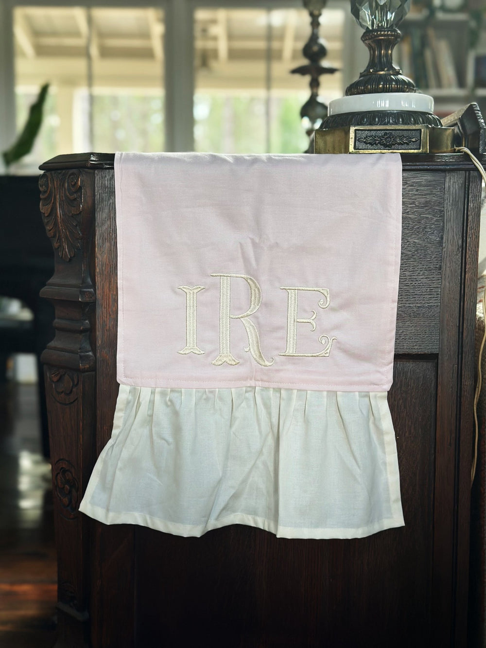 Monogrammed end of the baby pink runner draped over the side of an antique dresser, gathered ivory ruffle at the hem and a sunny window behind