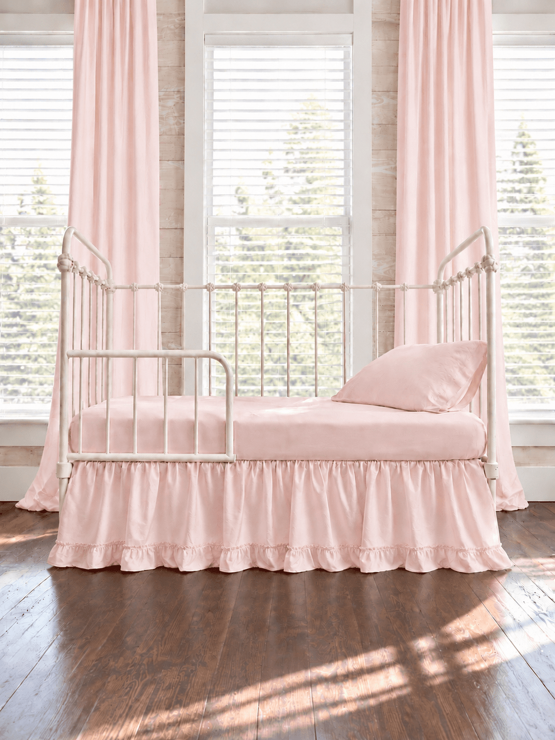 Baby pink ruffled toddler bed skirt with a gathered body and ruffled hem on a cream iron crib converted to a toddler bed, pink sheet, pillow and curtains - High Cotton Textile