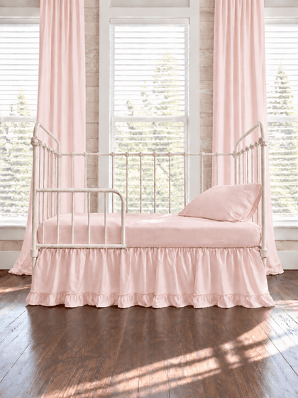 Baby pink ruffled toddler bed skirt with a gathered body and ruffled hem on a cream iron crib converted to a toddler bed, pink sheet, pillow and curtains - High Cotton Textile