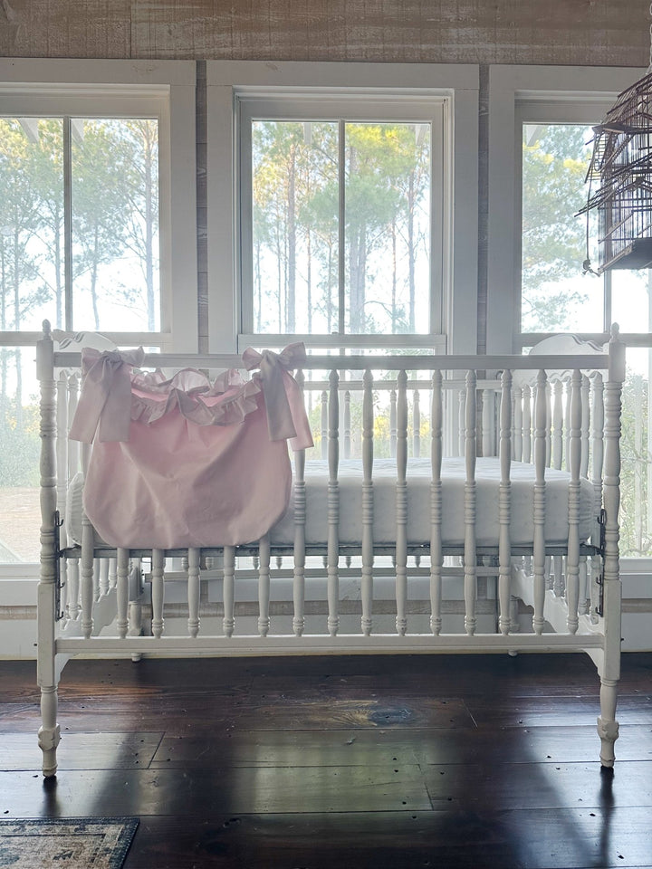 Baby pink ruffled caddy hanging on the outside of a white spindle crib beneath tall windows