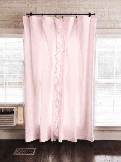 Baby pink ruffled nursery curtains hung on a black rod across a window, panels falling to dark wood floors - High Cotton Textile
