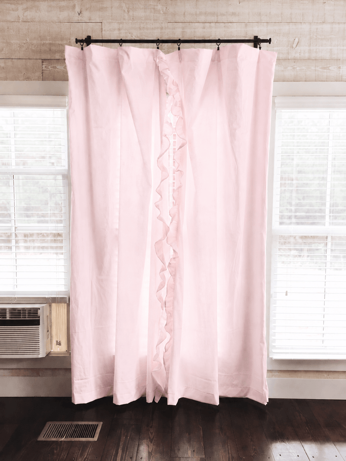 Baby pink ruffled nursery curtains hung on a black rod across a window, panels falling to dark wood floors - High Cotton Textile