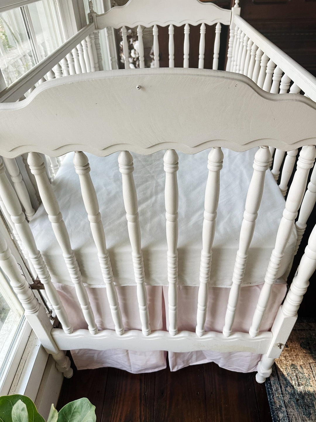 End view of the crib showing the baby pink tailored skirt's pleat under the scalloped footboard