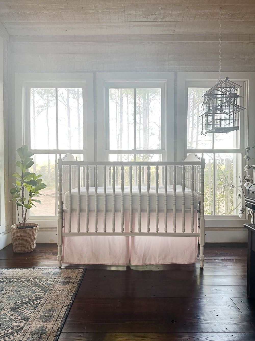 Nursery with a white spindle crib and baby pink tailored skirt in front of three tall windows, fiddle leaf fig and rug