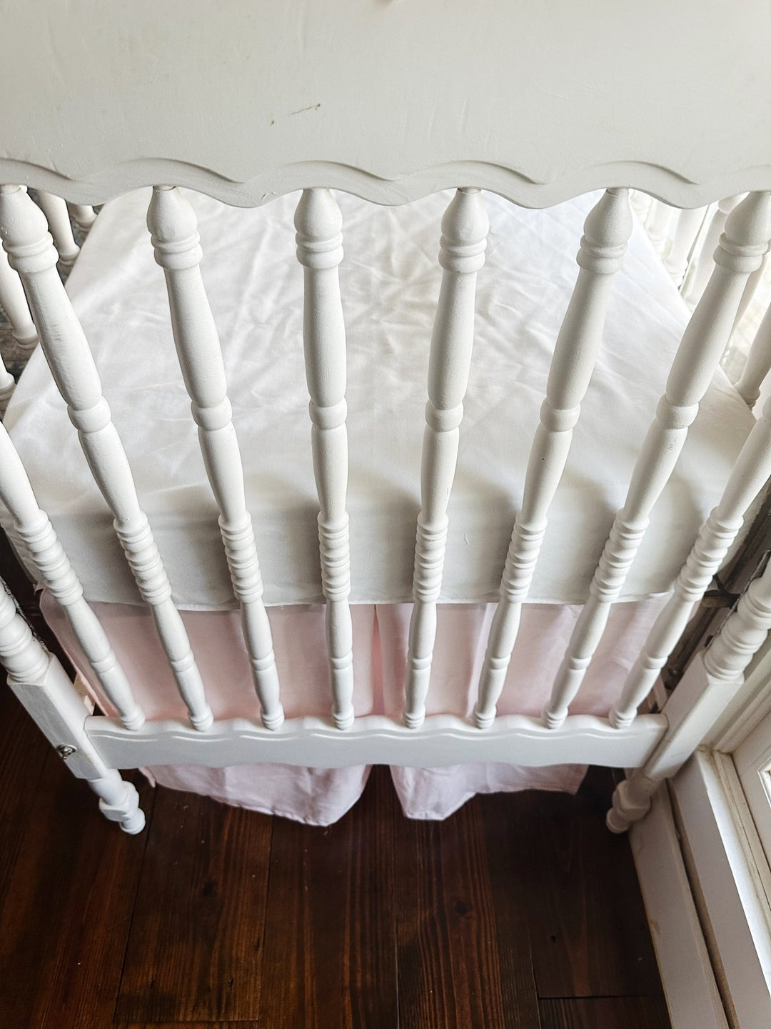 Short side of the baby pink tailored crib skirt with a center pleat, seen through the crib spindles