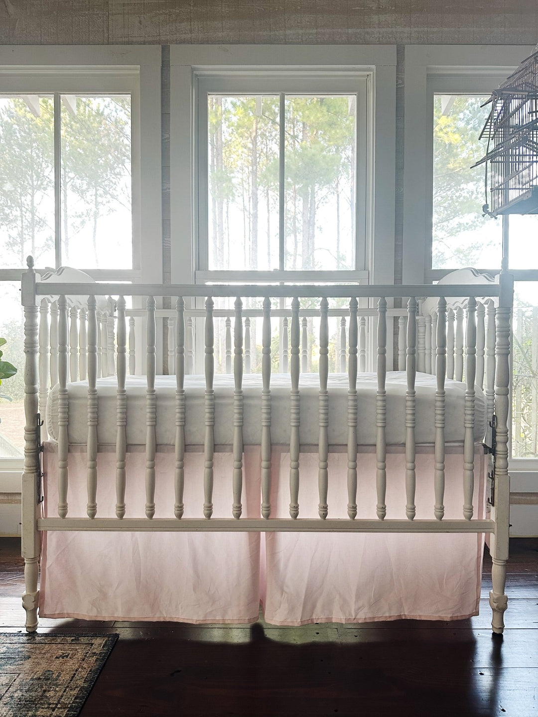 Baby pink tailored crib skirt with pleated center panels under an antique white spindle crib, front view