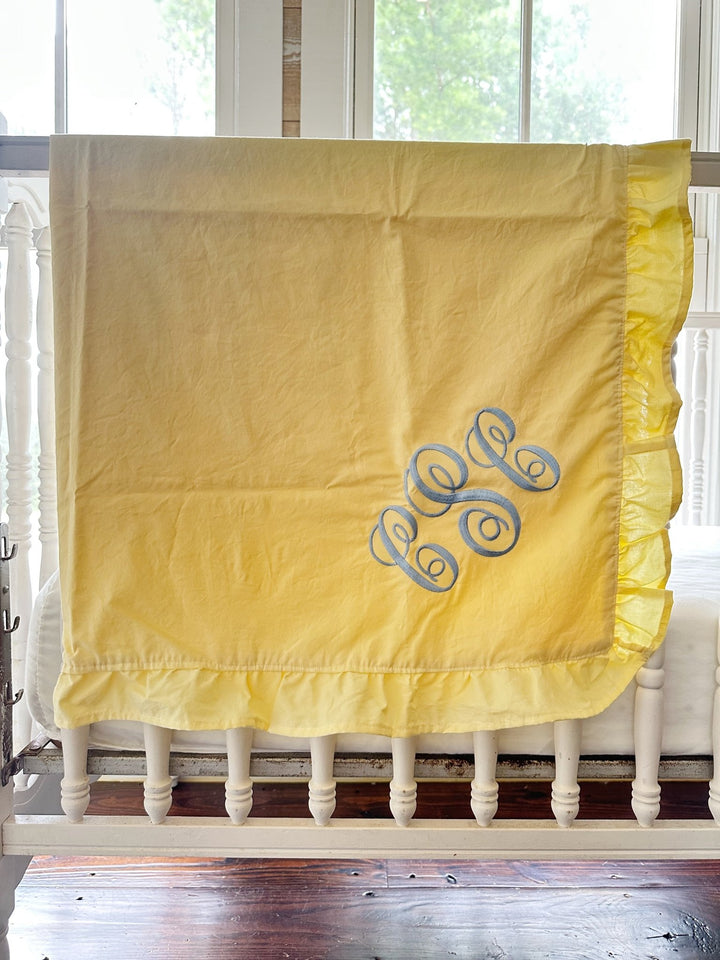 Baby yellow ruffled crib blanket with a blue monogram draped over a white spindle crib - High Cotton Textile