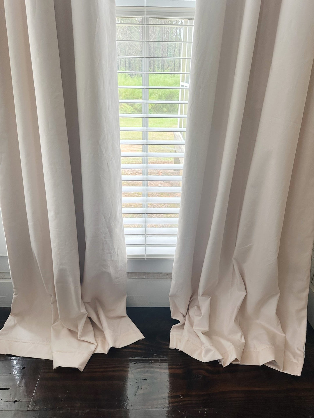 Bottom of two Ballet Slipper Pink blackout panels puddling softly on a dark wood floor below a window with white blinds