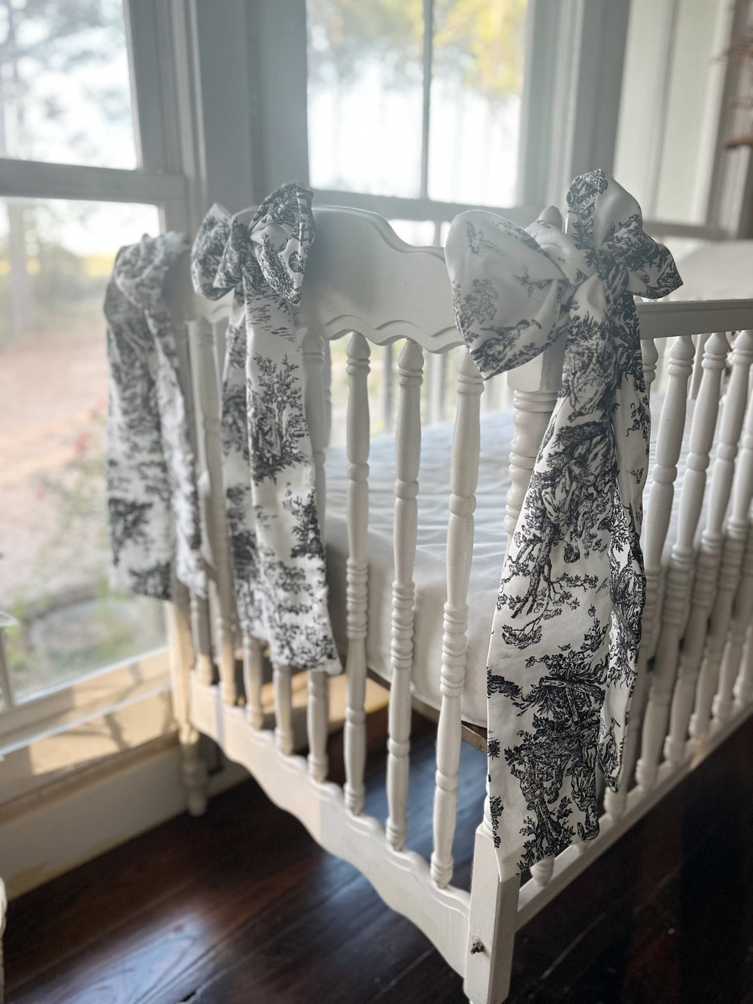 Three toile bows on the posts of an antique white crib in front of tall windows