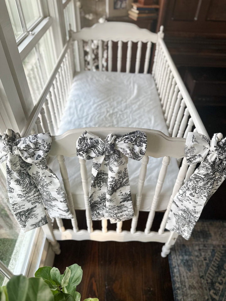 Two black toile bows on the end of a white crib beside a fiddle leaf fig