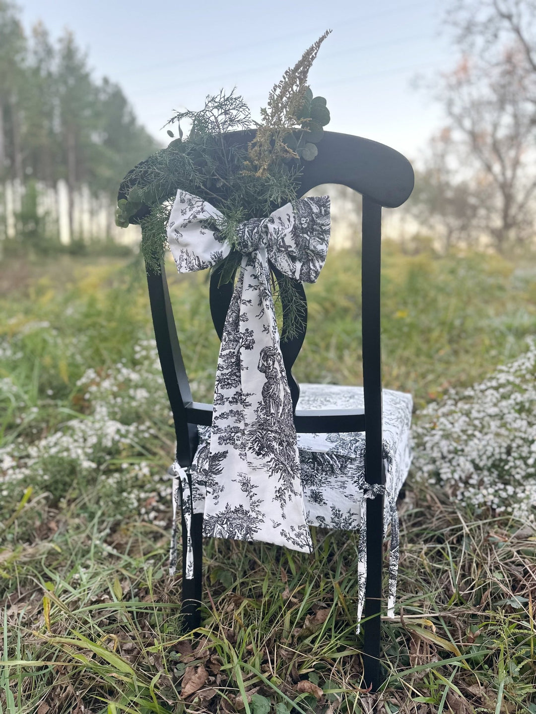 Black and white toile sash bow tied on a black dining chair outdoors with cedar and goldenrod tucked in