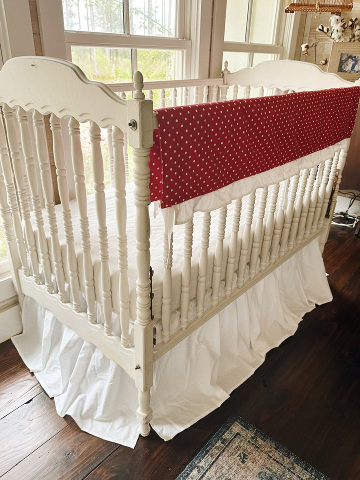 Corner view of the red polka dot rail cover running down the long rail of a white spindle crib