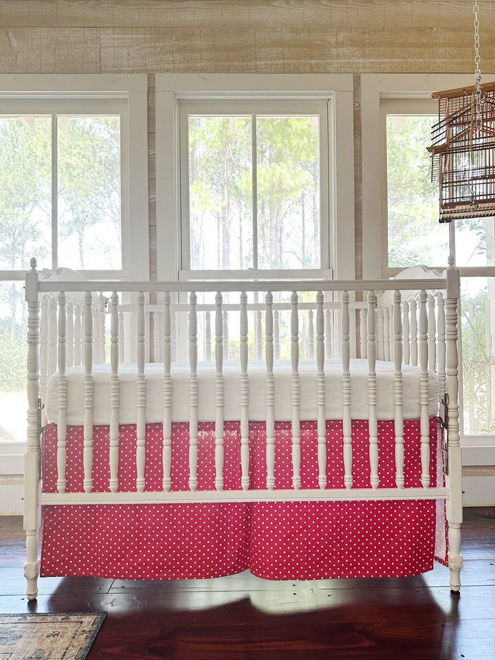 The dotted skirt straight on beneath a white crib, pine-paneled wall and three windows behind