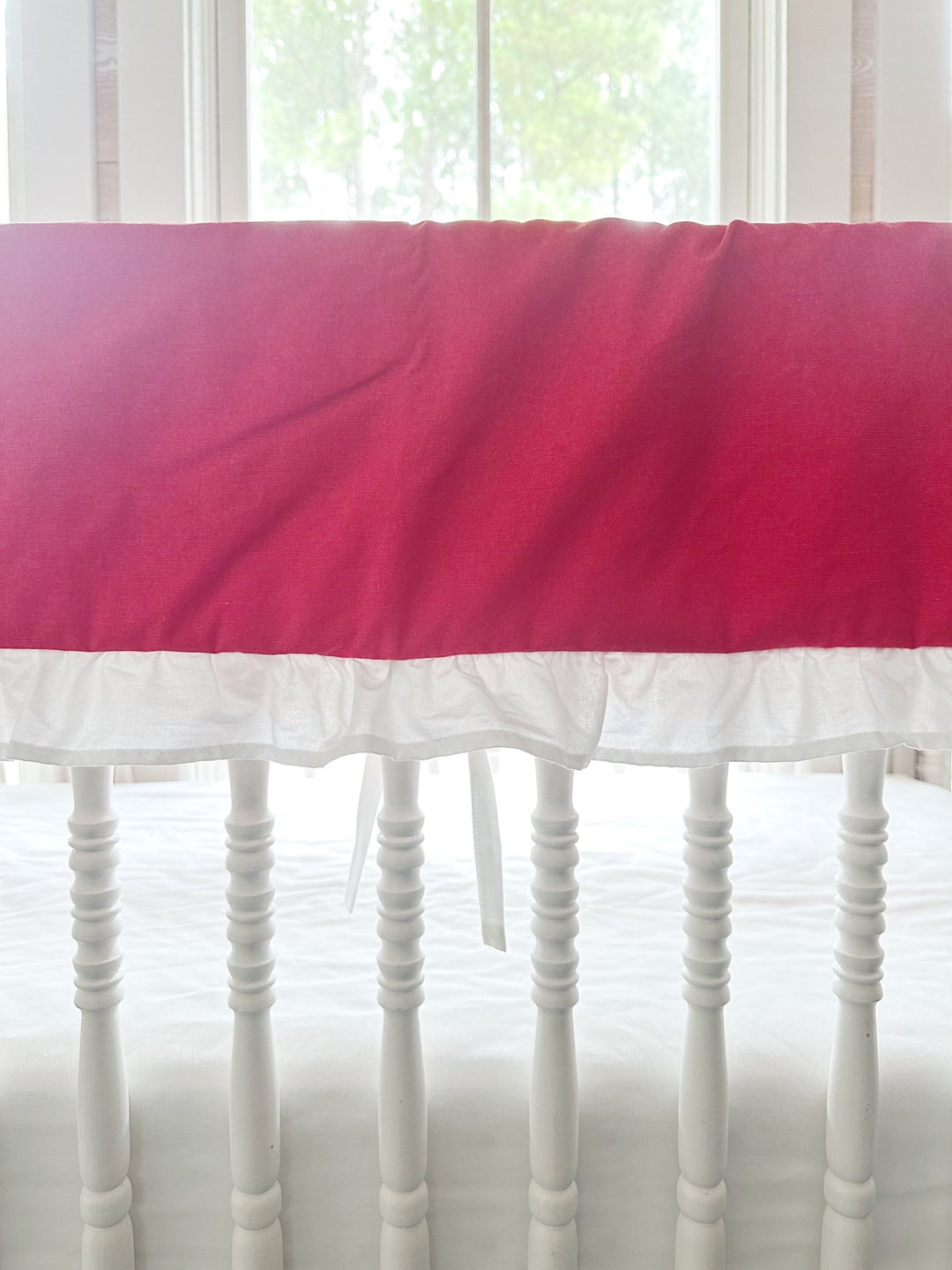 The red rail cover on the crib rail, showing the gathered white ruffle and slim white ties between the spindles