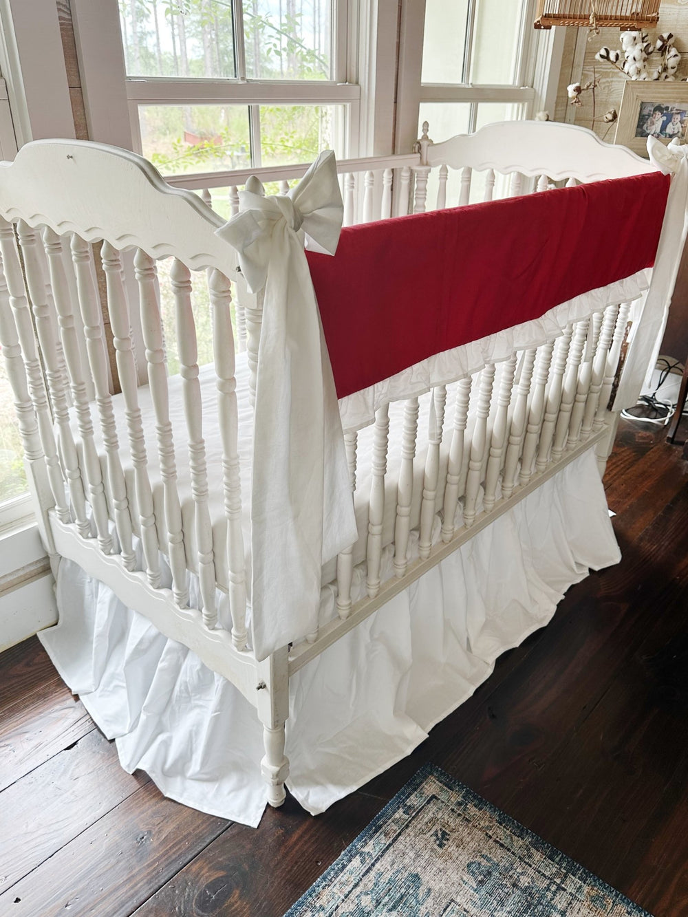 Corner view of the red rail cover running down the long crib rail with a large white bow on the near post