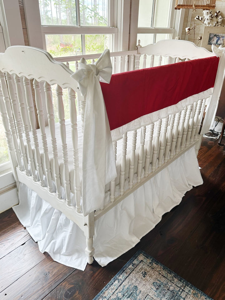 Corner view of the red rail cover running down the long crib rail with a large white bow on the near post