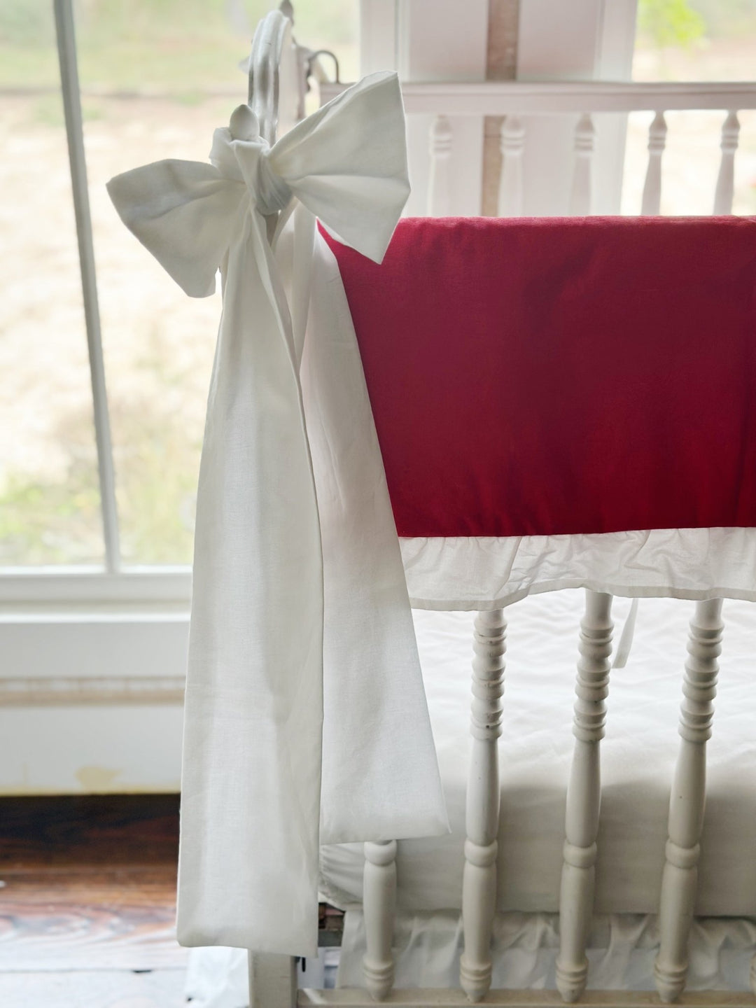 Large white bow knotted over the crib finial beside the red rail cover and its white ruffle